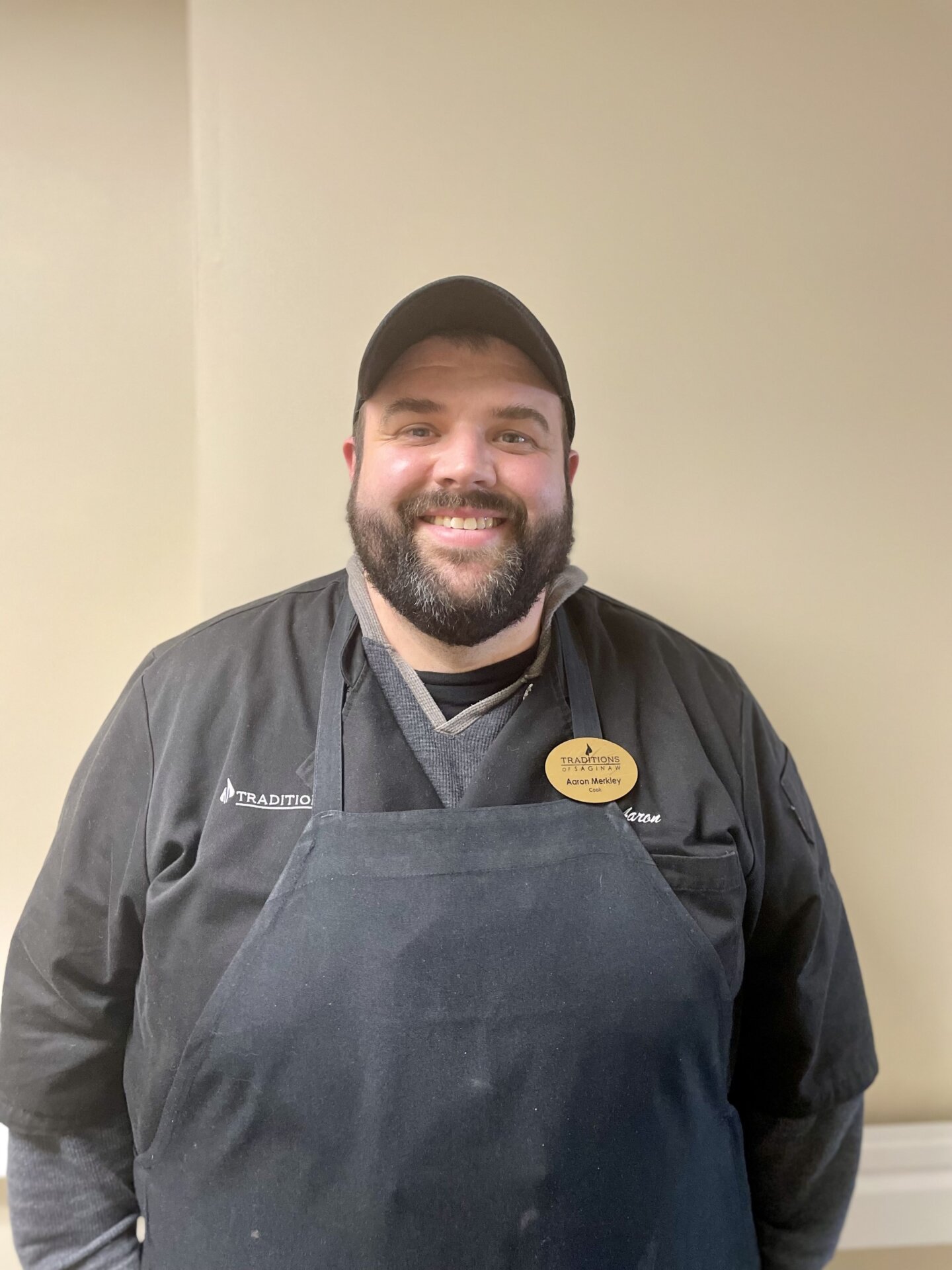 A male chef wearing a black hat and a blue apron with a gold name plate