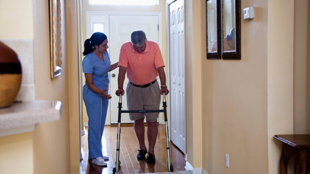 A caregiver helping an elderly man walk with his walker inside a house.