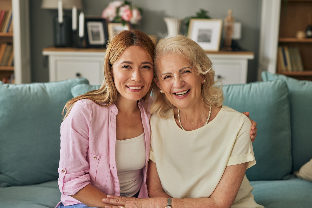 Mother and daughter smiling while sitting together on a couch, with the elderly mother wearing a cream blouse and the younger woman in a pink shirt, in a cozy living room setting.