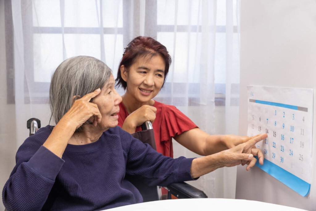 Female memory care patient sitting in a wheelchair as her female caregiver shows her what day it is on a calendar