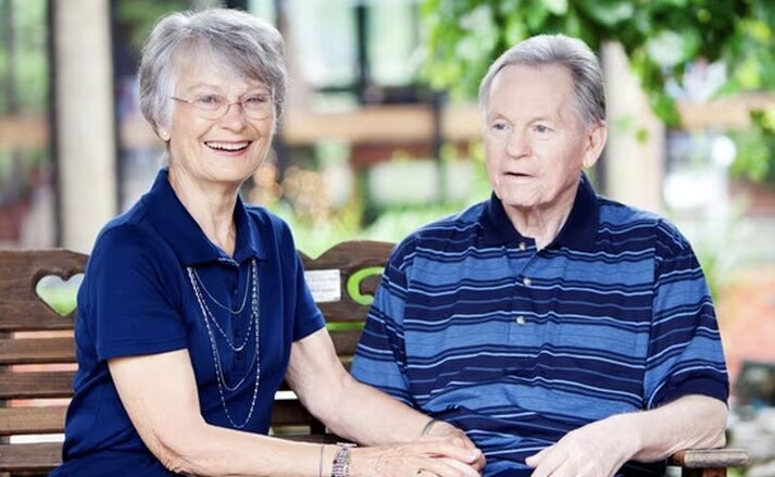 An elderly couple are sitting together on a bench while holding hands and enjoying the sunshine and nice weather. Traditions at Saginaw.