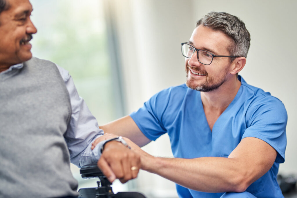 A male nurse kneels down next to a memory care resident. They are smiling and engaging in a friendly conversation. Traditions of Saginaw.