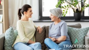 Photo of a woman with dark hair sitting on the couch across from her elderly mother as they have a conversation with the Traditions Management logo in the bottom right corner of the image